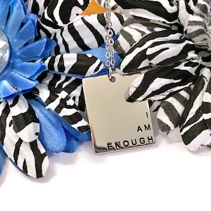 Può includere: Una collana d'argento con un ciondolo quadrato che recita "I AM ENOUGH". Il ciondolo è circondato da un tessuto a stampa zebrata blu e bianca.