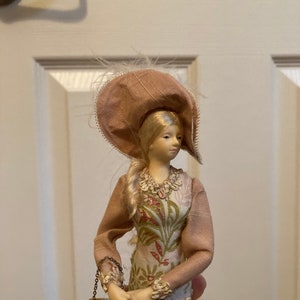 May include: A porcelain doll wearing a pink dress with floral print and a matching hat with feathers. The doll is holding a small, gold-trimmed purse.