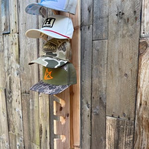 May include: A vertical wooden wall-mounted rack featuring four notched slots designed to hold baseball caps. Four distinct baseball caps are currently displayed on the rack, showcasing various colors, patterns, and logos including text.