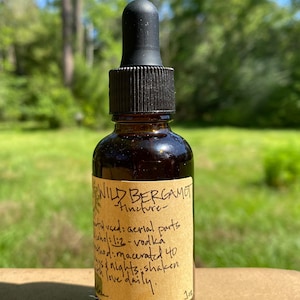 May include: A brown glass bottle with a dropper top and a label that reads "WILD BERGAMOT -Tincture-  plant veed: aerial parts alcohol: 1:2 - vodka method: macerated 40 days & nights, shaken with love daily. 1 oz"