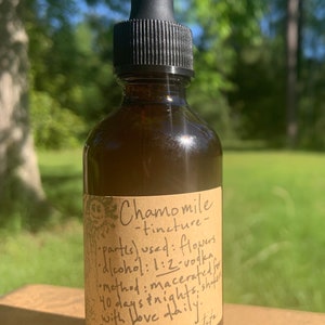May include: A brown glass bottle with a dropper top and a paper label that reads "Chamomile -tincture- parts) used: flowers dlcohol: 1:2-vodka Method: macerated 40 days Inights, shafer with love daily Matricaric recutipa"