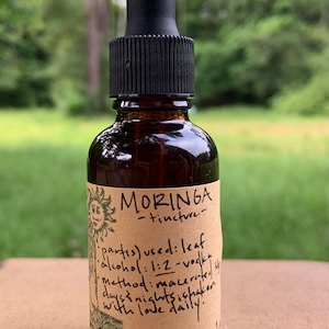 May include: A brown glass bottle with a black dropper top and a brown paper label. The label says "MORINGA -tincture- partes) used: leaf alkohol: 1:2-vok Method: macerated 4 days & nights shaken with love daily."