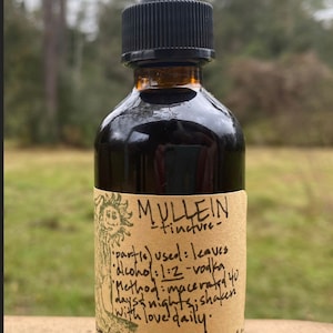 May include: A brown glass bottle of Mullein tincture with a black dropper. The label includes text about the ingredients and preparation method. The background is blurred, showing a natural outdoor setting.