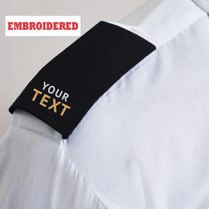 May include: Black shoulder epaulette with gold text reading "YOUR TEXT" on a white shirt. The word "EMBROIDERED" is in red text in the upper left corner. The epaulette is a rectangular shape.