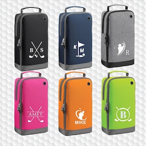 May include: A collection of golf shoe bags in various colors, including black, navy, gray, pink, orange, and green. Each bag features a handle, zipper, and a personalized golf-themed design with initials or a name. The bags have a gray base and trim.