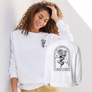 Puede incluir: Sudadera blanca con un diseño de rosa y serpiente negras. La parte delantera tiene un pequeño gráfico de rosa y serpiente. La parte trasera muestra un diseño más grande con una rosa, una serpiente y el texto "LIKE A ROSE IN A GRAVEYARD".
