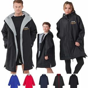 Puede incluir: Se muestran albornoces negros con capucha y forro polar gris. Los albornoces tienen una cremallera completa y el texto "YOUR TEXT" en dorado. Se muestran albornoces adicionales en azul, rojo, azul marino, negro con ribete rojo y negro.