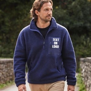 May include: A navy blue fleece pullover with a zip-up collar and a chest pocket. The pocket has the text "TEXT OR LOGO" printed on it in white. The pullover has a soft, textured appearance and is worn with khaki pants.