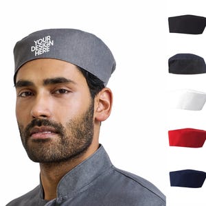 May include: A grey chef hat featuring the white text "YOUR DESIGN HERE" on the front. The hat is shown in a variety of colours including black, dark grey, white, red, and navy blue, displayed in a vertical column to the right of the main hat.