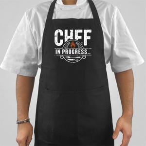 May include: Black apron with the text "CHEF IN PROGRESS" in white. The design includes a flame graphic and utensils. The apron has a large front pocket and is worn over a white chef's coat.