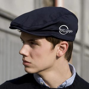 Puede incluir: Gorra plana azul marino con visera curva. La gorra tiene un logotipo circular blanco con el texto "Text Logo" y "your tagline here". La gorra se usa sobre una camisa de cuello azul claro y un suéter negro.