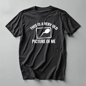 Peut inclure: T-shirt noir avec texte et graphisme blancs. Le texte dit "THIS IS A VERY OLD PICTURE OF ME" avec un dessin de spermatozoïde dans un rectangle blanc. Le t-shirt a un col rond et des manches courtes.