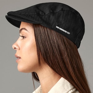 May include: A black flat cap with a rounded crown and a small, stiff peak. The cap features the white text "YOUR EMBROIDERED TEXT" embroidered on the side. The cap is made of a textured fabric.