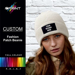 May include: A white knitted beanie with a black patch that says "YOUR DESIGN HERE". The beanie is being worn by a person with long brown hair.