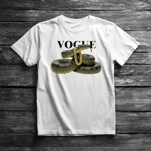 Puede incluir: Camiseta blanca con la palabra "VOGUE" en negro sobre un gráfico de una serpiente verde y negra con una cadena dorada. La serpiente está enrollada y centrada en la camiseta.