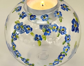May include: A clear glass globe candle holder with a tealight candle inside. The globe is decorated with hand-painted blue forget-me-not flowers.