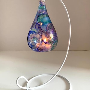 May include: A decorative glass candle holder in a teardrop shape, with blue, purple, and teal abstract patterns. It hangs from a white metal stand, with a lit candle inside. The stand is made of metal.