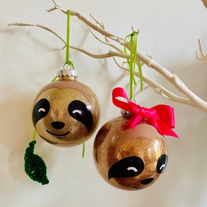 May include: Two gold glitter Christmas baubles decorated with sloth faces, suspended from a white branch. One has a green ribbon, the other a pink bow. The sloths have black eyes and noses, and a light brown face.