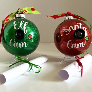 Może przedstawiać: Dwie ozdoby choinkowe, jedna zielona, a druga czerwona, z brokatem i napisami "Elf Cam" i "Santa Cam". Każda ozdoba ma mały obiektyw kamery i stoi na podstawce z zwiniętym kawałkiem papieru związanym wstążką.