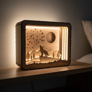 May include: A wooden night light with a layered design featuring a howling wolf silhouette, trees, mountains, a moon, and stars. The rectangular light is illuminated from within, creating a warm glow. It sits on a wooden nightstand.