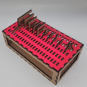 May include: A wooden organizer with a bright pink interior and laser-cut grid pattern. The organizer has several slots holding small, brown wooden cutouts of various shapes. The box is rectangular with a natural wood finish.