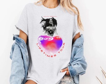 Puede incluir: Una camiseta blanca con un gráfico de Katy Perry en una pose de superhéroe sosteniendo un corazón con el texto "KP" en su interior. El corazón está rodeado por un efecto de acuarela rosa y azul y el texto "LIFETIME TOUR" está escrito alrededor del corazón.