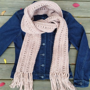 Puede incluir: Una bufanda de crochet rosa claro con flecos en los extremos. La bufanda está drapeada sobre una chaqueta vaquera. El texto "PDF PATTERN" está en la esquina inferior izquierda de la imagen y el texto "ESTHER'S HANDS" está en la esquina inferior derecha de la imagen.