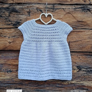 May include: A light purple crochet baby dress with short sleeves hanging on a white heart-shaped hanger against a wooden background. The dress has a simple design with a crocheted pattern. The text "PDF PATTERN" and "ESTIER'S HANDS" are visible at the bottom of the image.