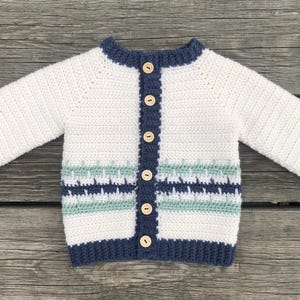 Könnte beinhalten: Weißer Baby-Strickcardigan mit langen Ärmeln, marineblauen Abschlüssen an Kragen, Bündchen und Saum. Die Vorderseite hat einen marineblauen Knopfverschluss und ein dekoratives Band mit blauen und grünen geometrischen Mustern.