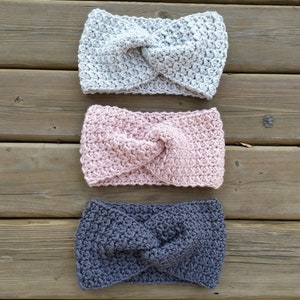 May include: Three crocheted headbands in gray, pink, and dark gray. The headbands are made with a textured stitch and have a twisted knot in the center. The headbands are displayed on a wooden surface. PDF PATTERN ESTHER'S HANDS
