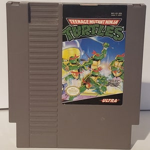 May include: A Teenage Mutant Ninja Turtles video game cartridge for the Nintendo Entertainment System (NES). The cartridge is grey with a black label featuring the Teenage Mutant Ninja Turtles logo and a scene of the four turtles fighting. The label also includes the text "NES-U2-USA" and "Made in Japan".