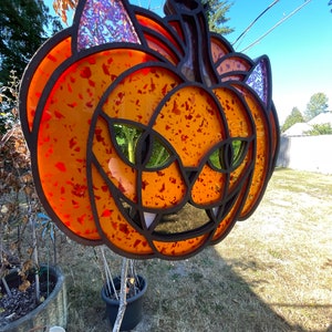 May include: A wooden pumpkin with a cat face design. The pumpkin is painted orange with red and black accents. The cat's eyes are green and the ears are purple.