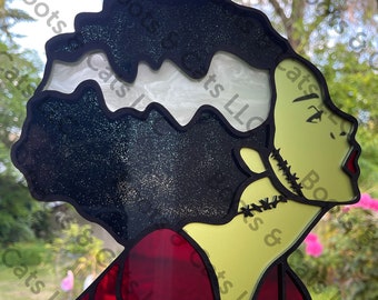 May include: A stained glass ornament featuring a green-skinned woman with black hair and a red dress. The woman has stitches on her neck and a white scar on her forehead. The ornament is made of glass and has a glittery finish.