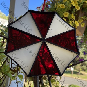 May include: A red and white stained glass ornament shaped like the Umbrella Corporation logo from the Resident Evil video game series. The ornament is hanging from a branch.