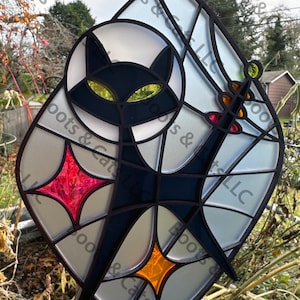 May include: Stained glass suncatcher featuring a black cat with yellow eyes, a sword, and geometric shapes. The design incorporates red and orange starbursts and a clear background. The suncatcher is suspended by a black cord.