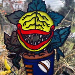 May include: A stained glass window hanging ornament featuring a cartoon plant monster with a toothy grin, red lips, and yellow and green leaves. The plant is in a blue pot with brown stripes.