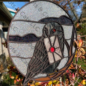 May include: A round, silver glitter-covered ornament with a black silhouette of a bird carrying a red and white envelope. The ornament has a brown wooden frame.