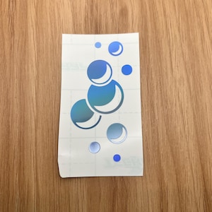 May include: A sticker featuring a design of blue bubbles in various sizes. The bubbles have a gradient effect, transitioning from a darker blue to a lighter, almost white color. The sticker is on a white backing.