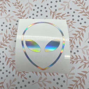 May include: A white sticker with a holographic alien head design. The alien has a large, round head with two large, oval eyes.