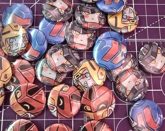 May include: A collection of circular pin-back buttons featuring stylized illustrations of robot faces in various colors including red, blue, yellow, and grey. Each button displays a unique character design with expressive eyes and metallic-looking armor plating.