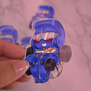 May include: A die-cut vinyl sticker featuring a chibi-style blue robot character with a visor, blushing cheeks, and a wide smile. The robot has black wheels on its sides and is holding a small brown animal figure against its chest.