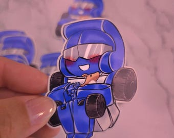 May include: A die-cut vinyl sticker featuring a chibi-style blue robot character with a visor, blushing cheeks, and a wide smile. The robot has black wheels on its sides and is holding a small brown animal figure against its chest.