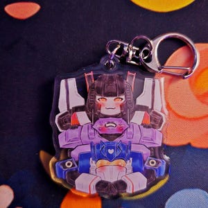 May include: A clear acrylic keychain featuring a cartoon illustration of a robot character in purple, blue, and white. The keychain has a silver metal clasp and ring. The background is a dark blue fabric with floral patterns.