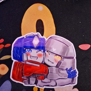 Op de afbeelding: Een sticker met twee cartoonrobots die elkaar omhelzen. Eén is voornamelijk rood en blauw, de ander zilver. Beide hebben lachende gezichten en hartjes. De achtergrond is zwart met een gele vorm en bloemmotieven.