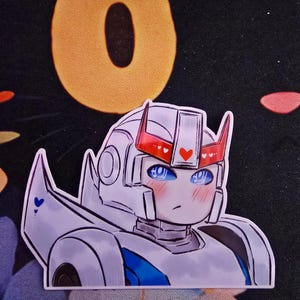 May include: A sticker featuring a cartoon robot character with a white and blue color scheme. The robot has red accents, including hearts, and blushing cheeks. The background is black with a large orange eye.