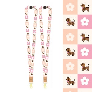 May include: Two lanyards featuring a repeating pattern of brown dachshunds wearing pink bandanas and white flowers on a checkered background of light pink and peach squares. Each lanyard includes a black plastic buckle and a gold metal swivel hook.