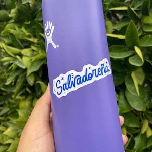 May include: A purple reusable water bottle with a white hand logo and a white sticker that says "Salvadoreña" in blue lettering.