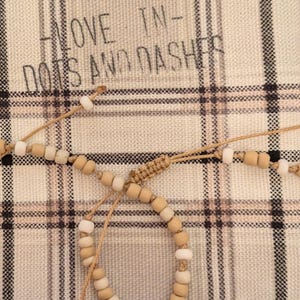 Puede incluir: Dos pulseras de cuentas con cuentas de madera y blancas descansan sobre una bolsa a cuadros. La bolsa tiene el texto "LOVE IN - DOTS AND DASHES" impreso. Las pulseras están atadas con un cordón de color natural.