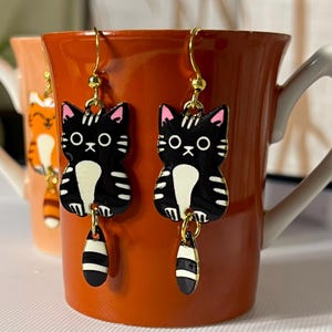 May include: A pair of black cat earrings with gold-colored hooks. The earrings feature a black cat design with white and pink accents, and a dangling striped charm. The earrings are displayed on a red mug.