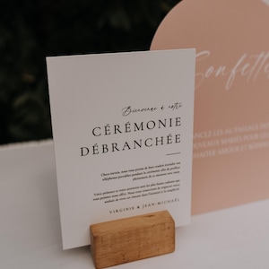 Peut inclure: Une carte blanche avec du texte noir sur un support en bois. Le texte indique "Bienvence à notre CÉRÉMONIE DÉBRANCHÉE". La carte est posée sur une surface blanche.